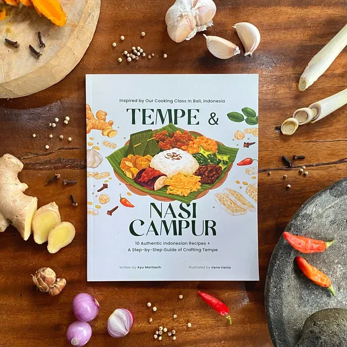 The cookbook Tempe and Nasi Campur by Ayu Martiasih and Irene Vania on a wooden surface surrounded by fresh ingredients